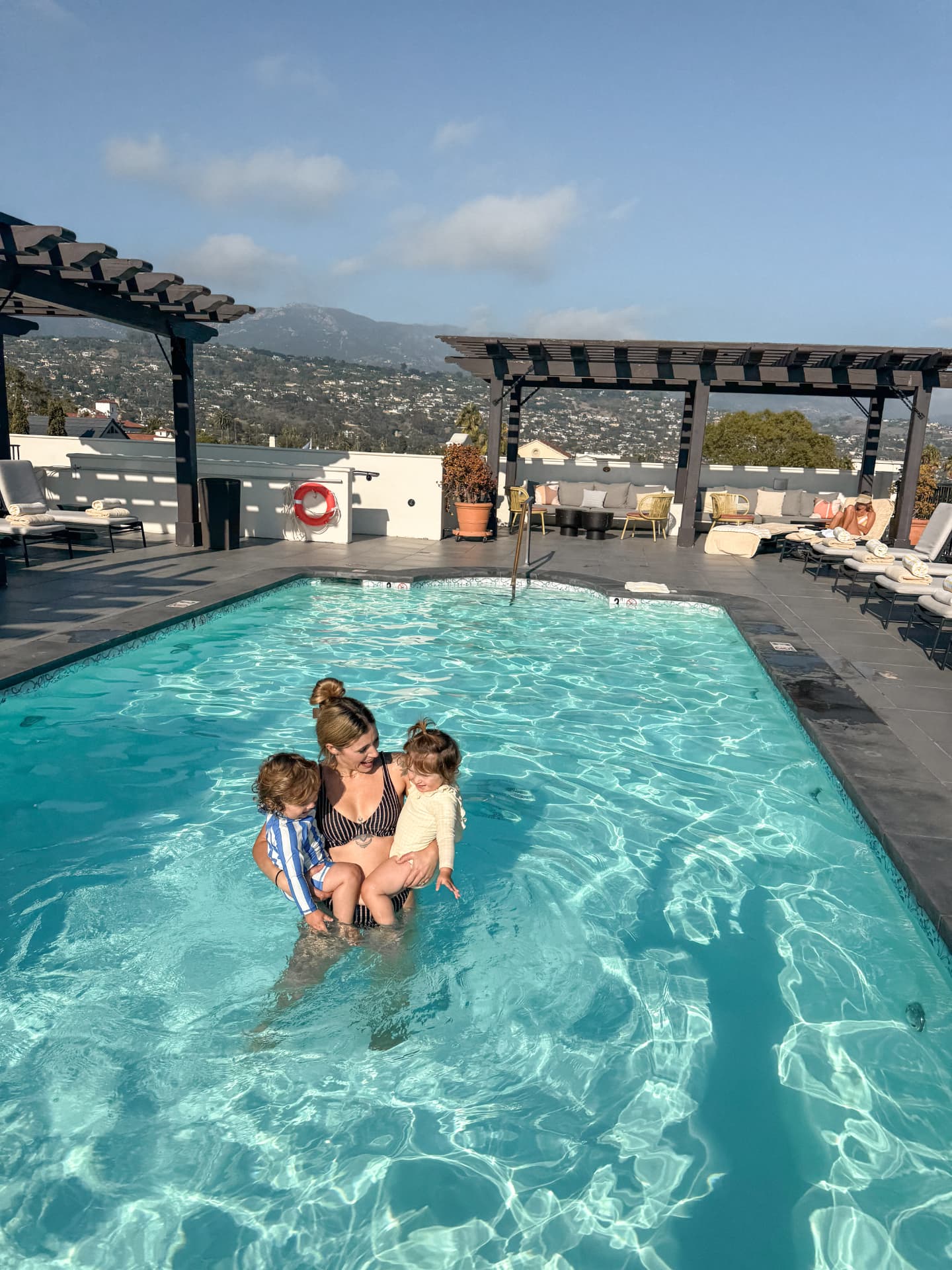 Linda standing in the rooftop pool at the Kimpton Canary Hotel holding August and Amelia, with pergolas, loungers and the Santa Barbara foothills behind them