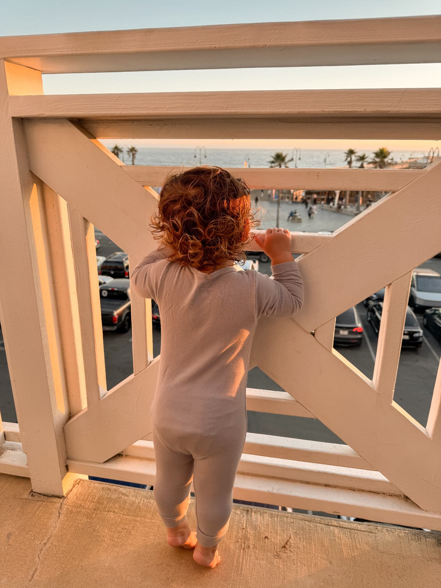 A small child standing at a wooden balcony railing in golden evening light, looking out to the ocean and pier beyond